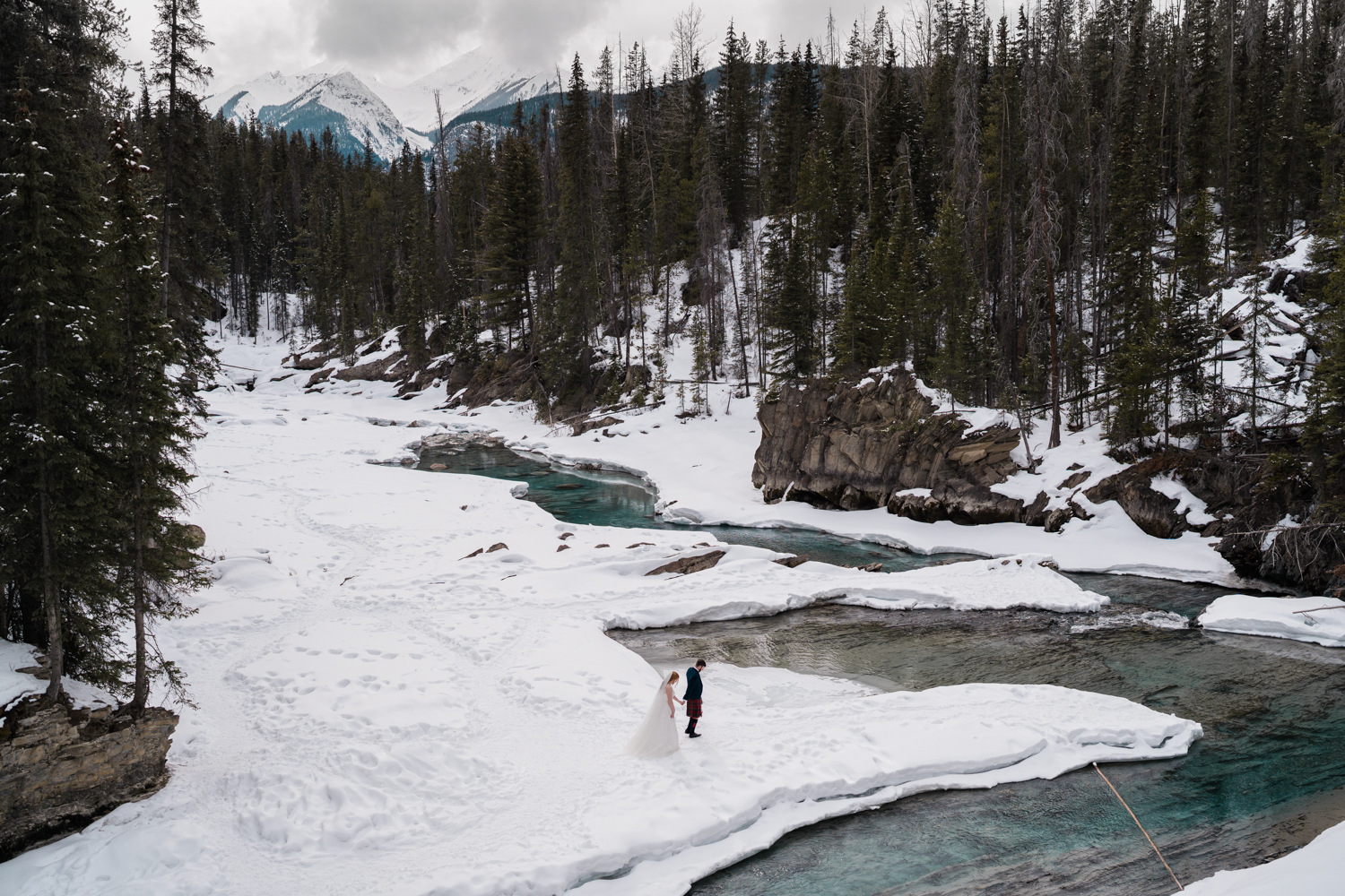 groom leads bride along the shorelines of the Kicking Horse River which snakes around behind them under snowy peaks for their Emerald Lake elopement