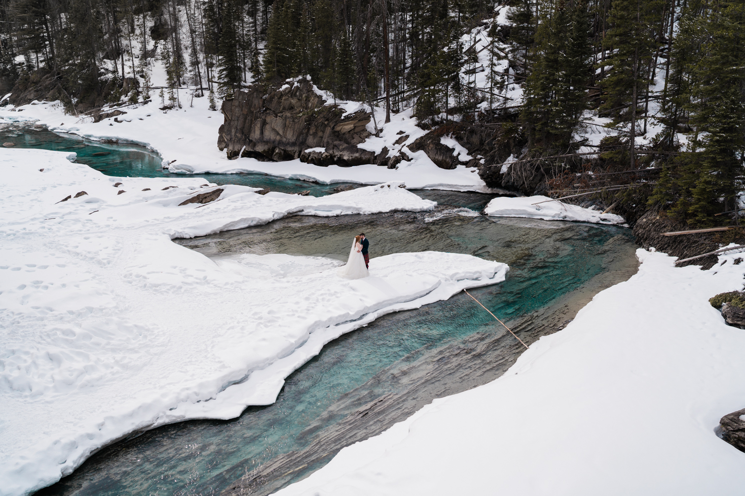 Bride and groom snuggle on a snowy outcrop in the Kicking Horse River at the natural bridge