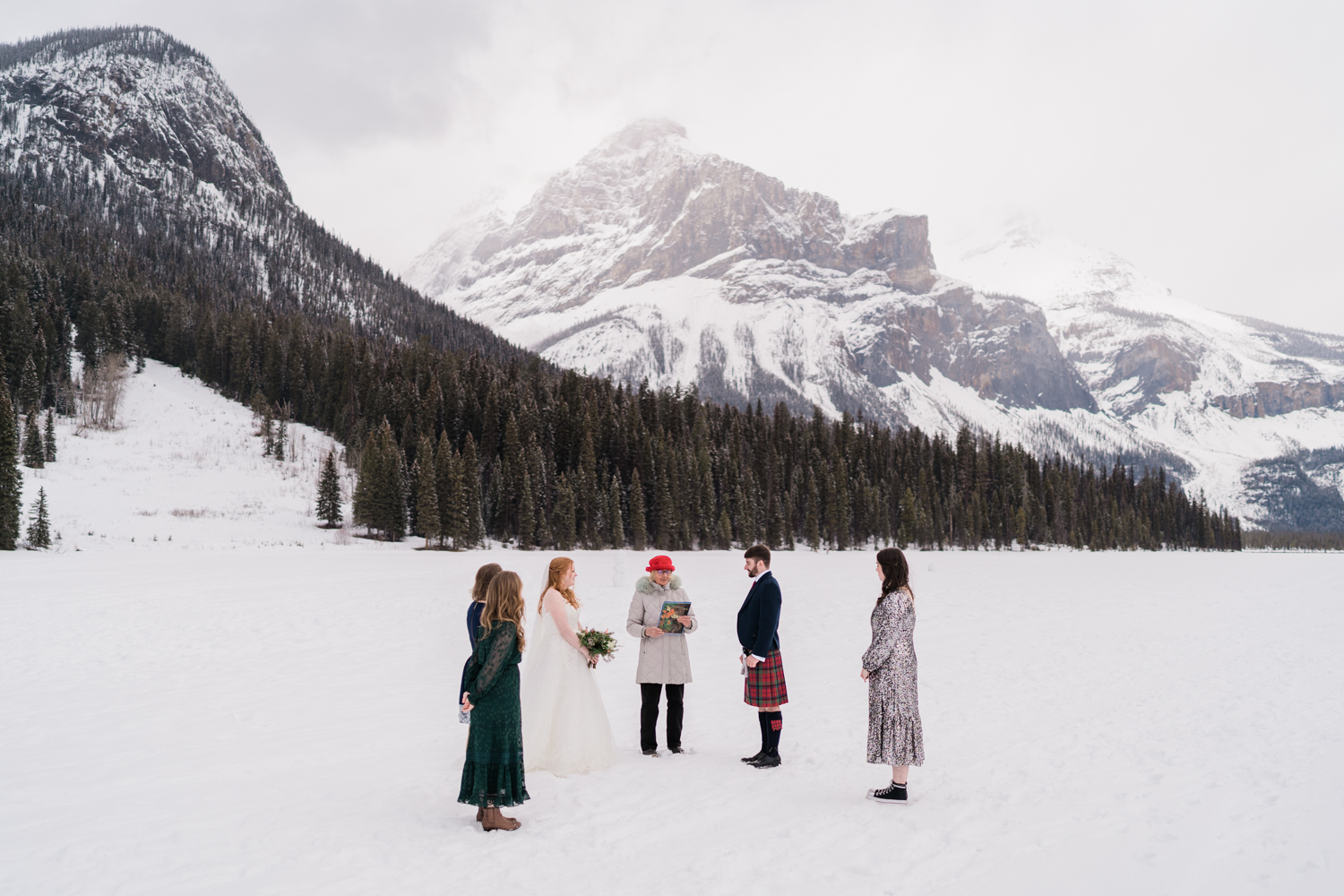 a bride and groom with three friends stand together on a frozen emerald lake during a wedding ceremony