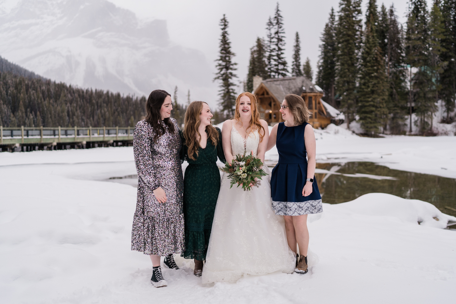 bride smiles holding bouquet with three friends around her in the snow infront of Emerald Lake lodge