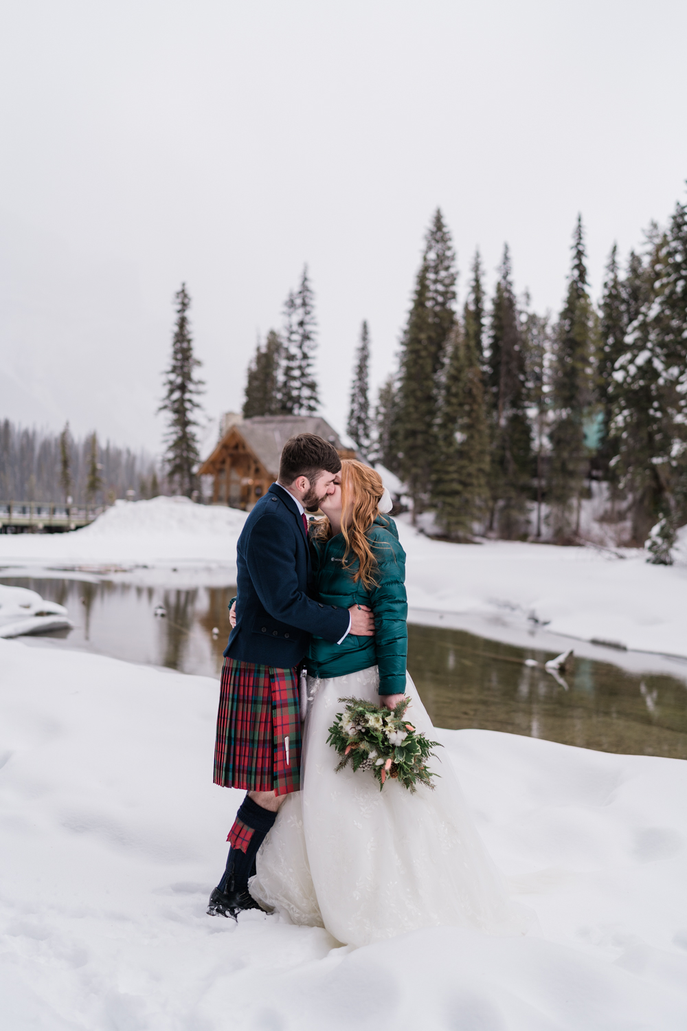 bride in green puffy and groom in kilt kiss while standing on the snow infront of Emerald Lake Lodge
