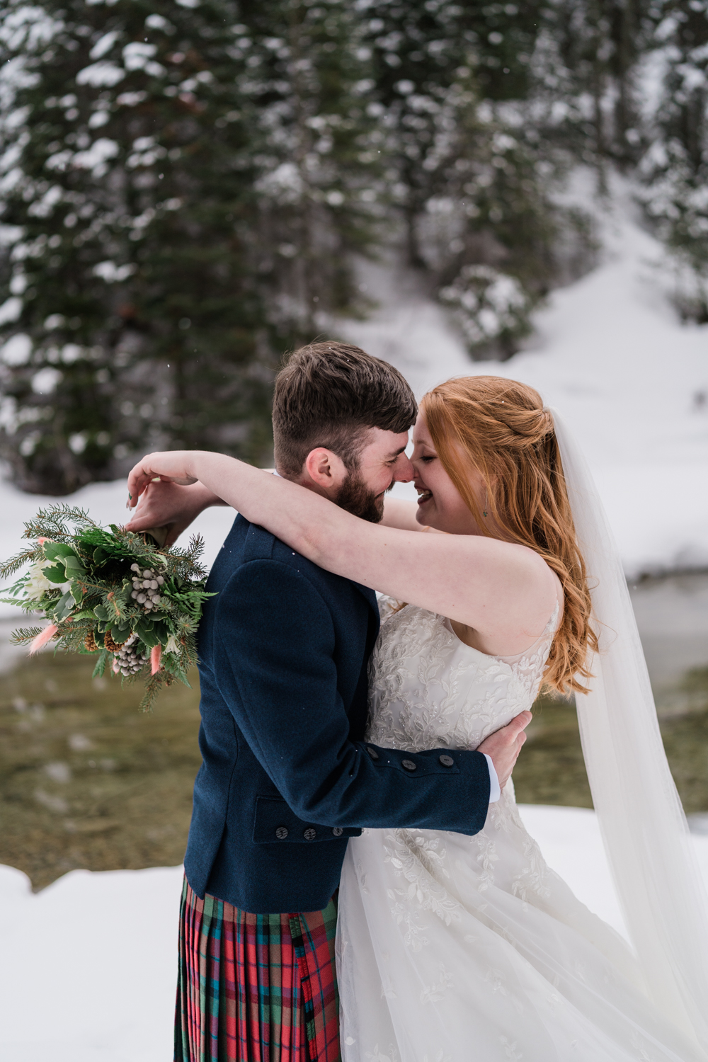 close up of bride and groom smiling at each other while she holds flowers and the snow lightly falls while the stand infront of a river