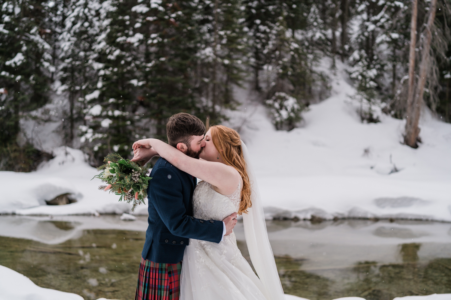 bride and groom kiss infront of open water in Yoho National Park while the snow lightly falls