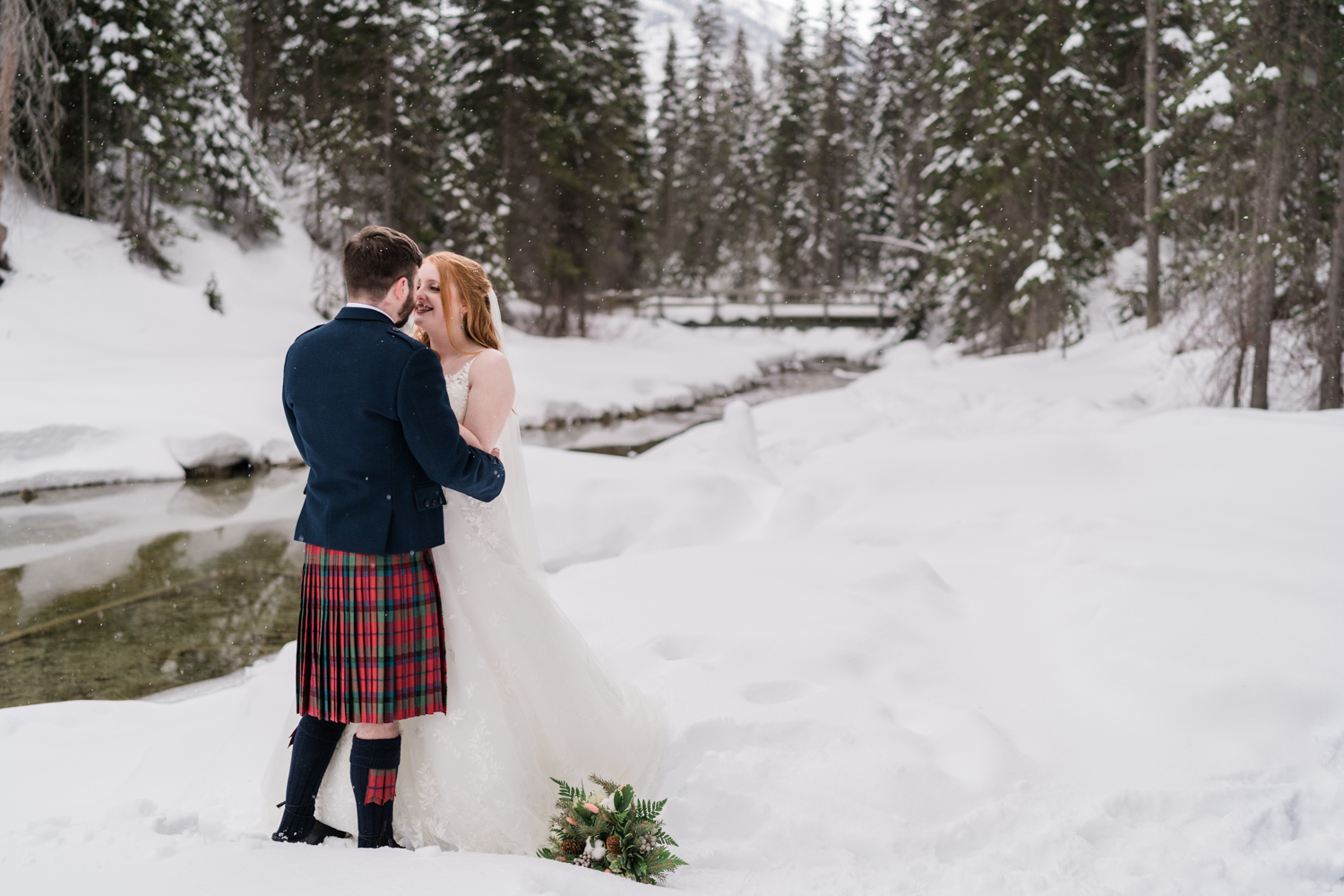 bride and groom face each other while the snow falls lightly with a snowy forest and bridge in the background