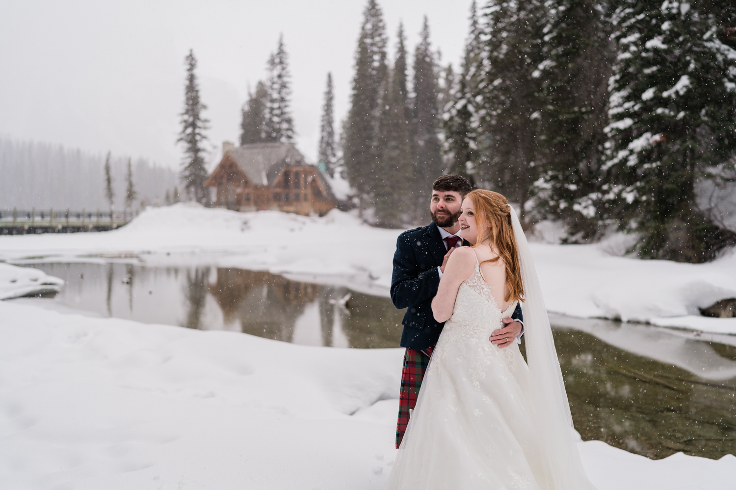the snow falls on a bride and groom as they stand and look off to the left infront of Emerald Lake Lodge during a winter elopement