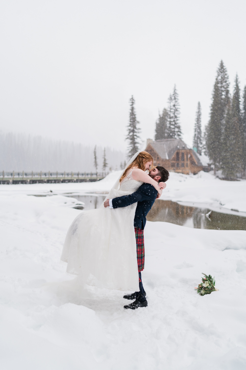 Groom in kilt holds up bride as the snow falls around them in front of the Emerald Lake Lodge during a winter elopement