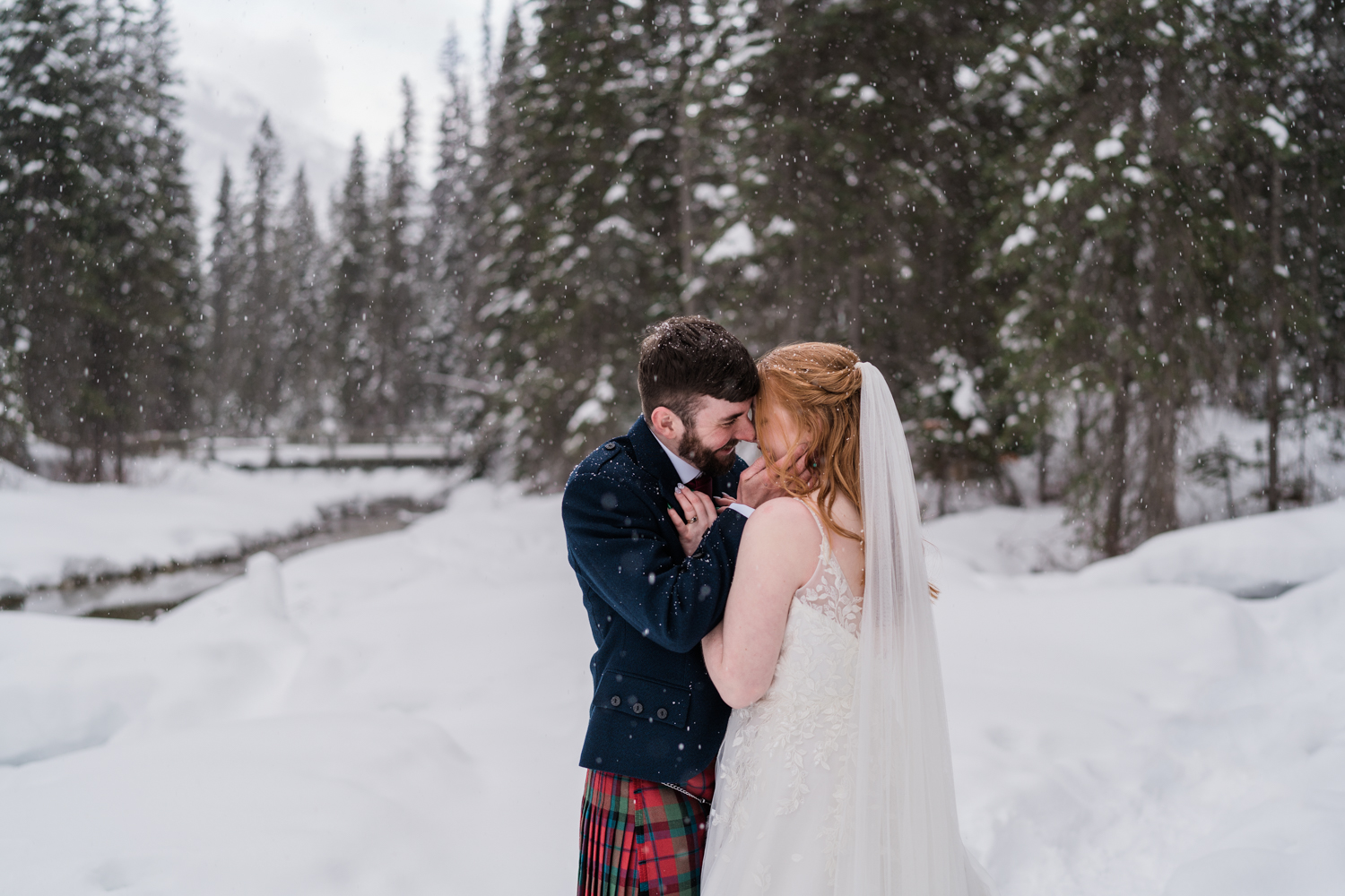 A bride and groom snuggle up with the snow falling down in the forest during an Emerald Lake elopement