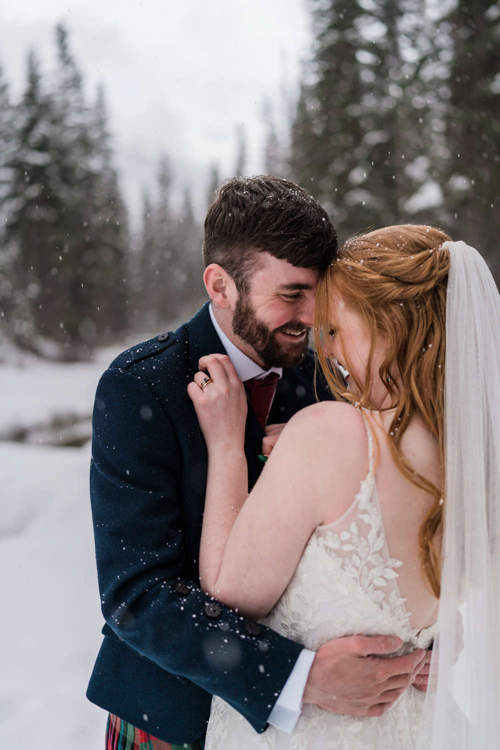 Bride with green nails snuggles up with groom as the snow falls down around them and they laugh together during an Emerald Lake elopement