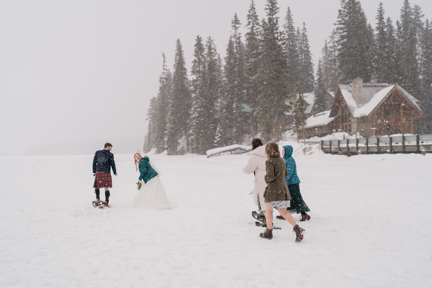 Bride and groom snowshoe with three friends walking behind them in front of Emerald Lake Lodge in a snowstorm