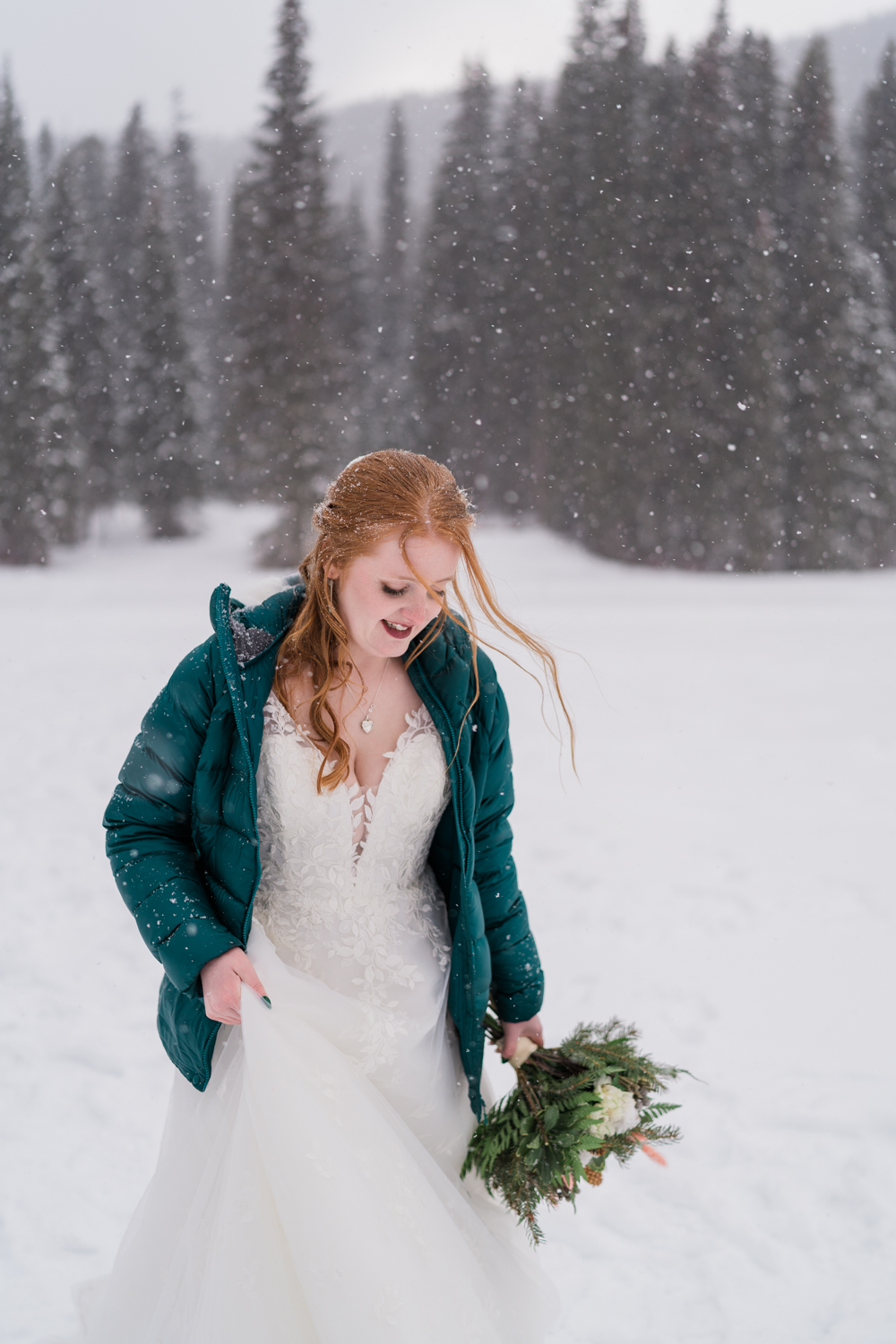 A red headed bride in a green puffy jacket walks looking down while holding bouquet in a snowstorm during Emerald Lake elopement