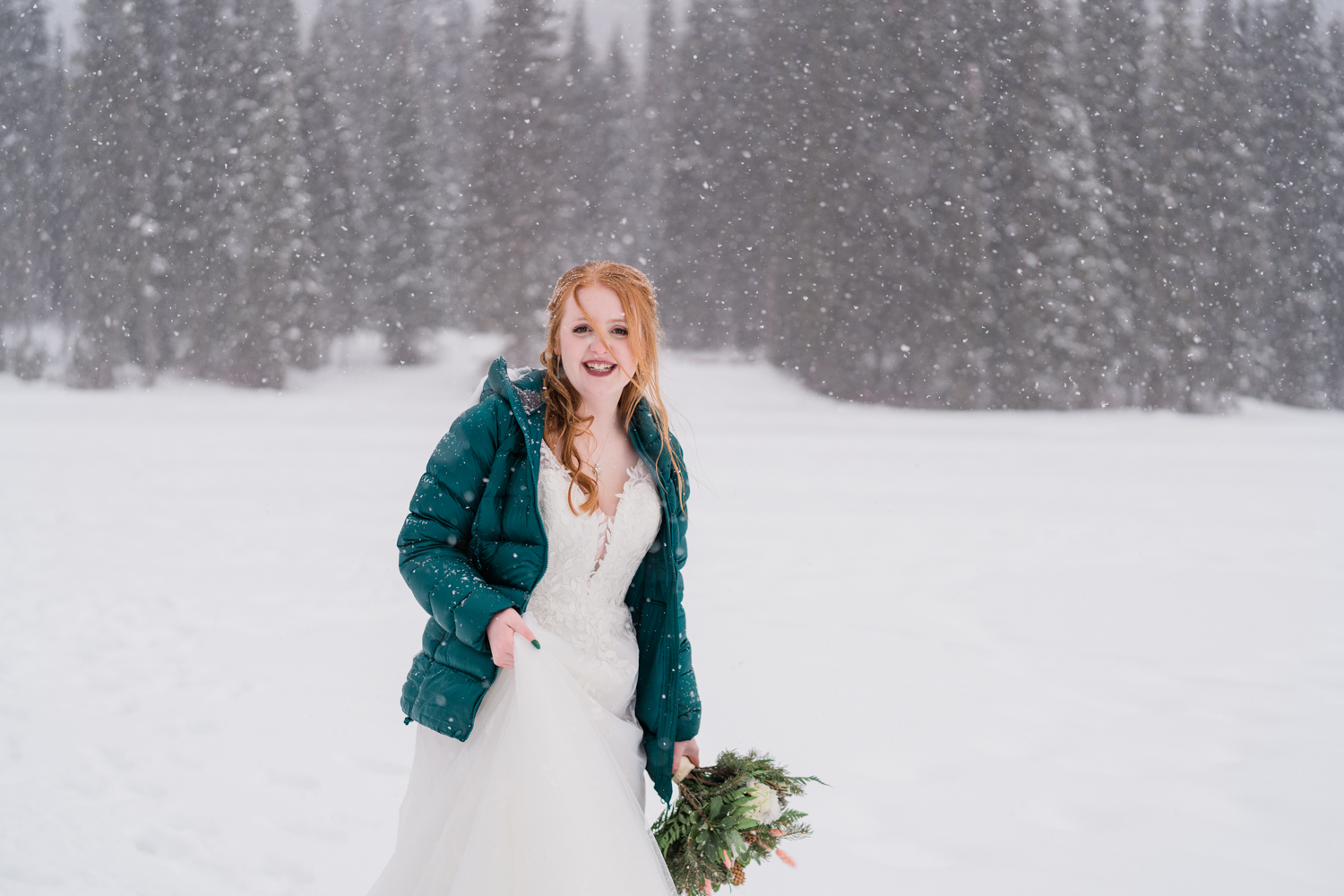A red headed bride in a green puffy jacket smiles at camera while holding bouquet in a snowstorm during Emerald Lake elopement