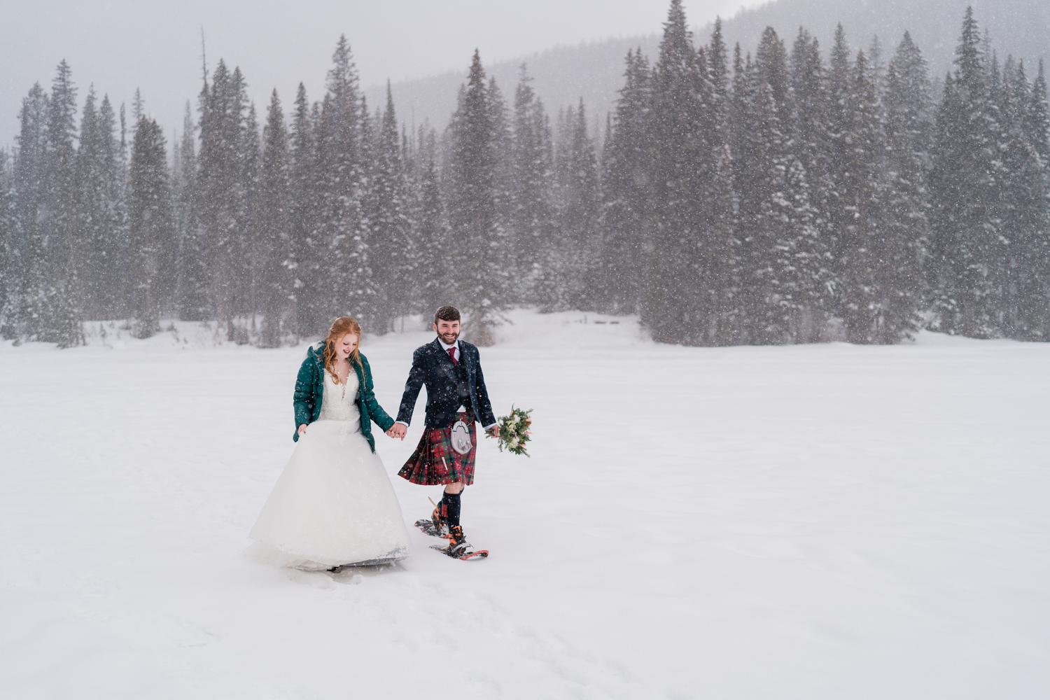 A red headed bride in a green puffy jacket snowshoes with groom in a kilt in a snowstorm during Emerald Lake elopement