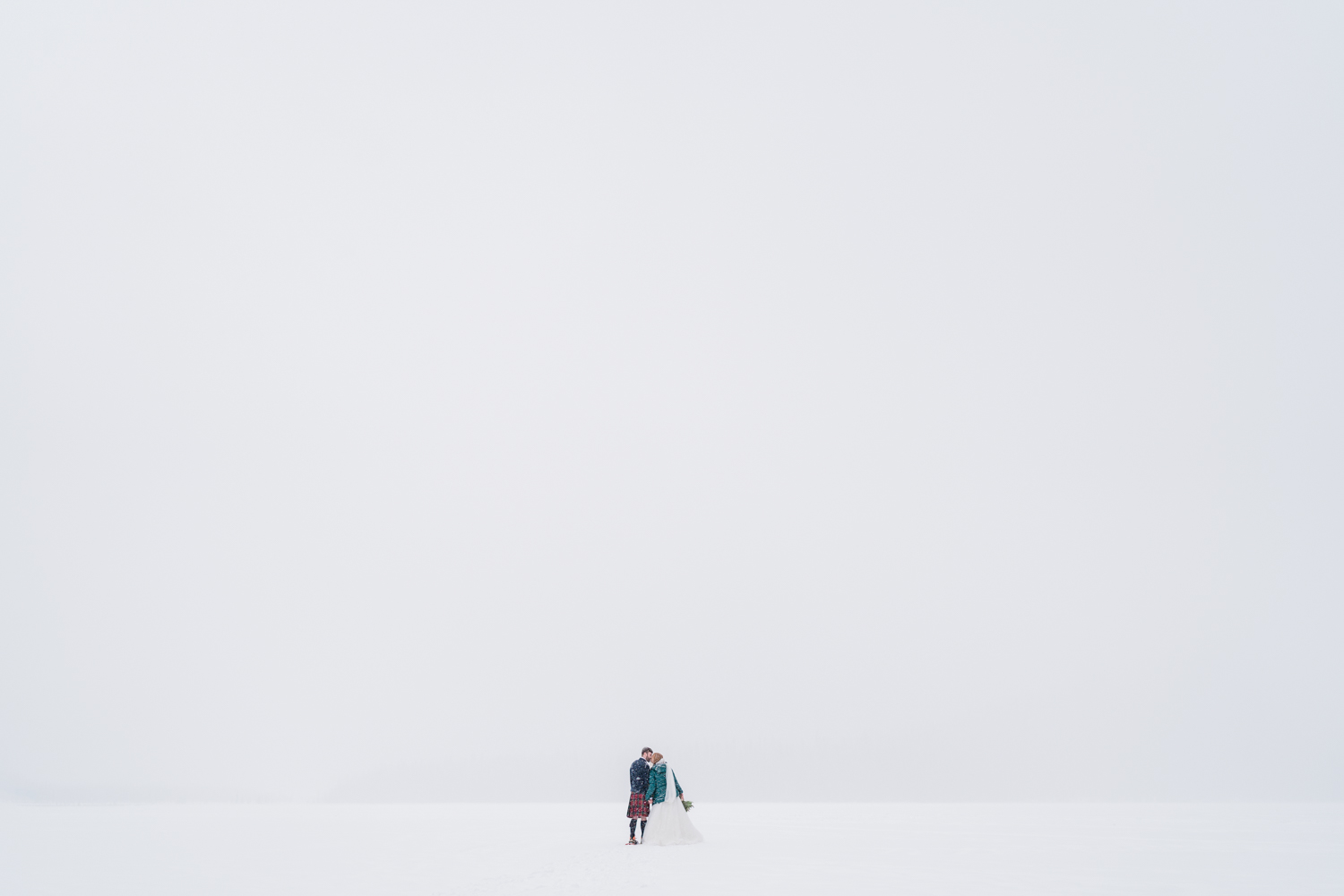 a bride in a green puffy jacket and a groom in a kilt kiss with a bunch of negative space above them from a whiteout in a snowstorm during an Emerald Lake elopement