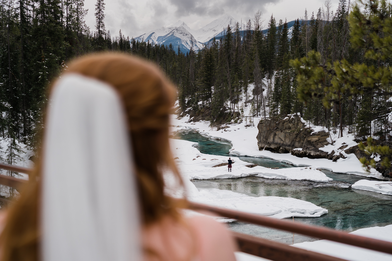back of brides head blurred out looking down at groom standing along the shore lines of the Kicking Horse River during their Emerald Lake elopement