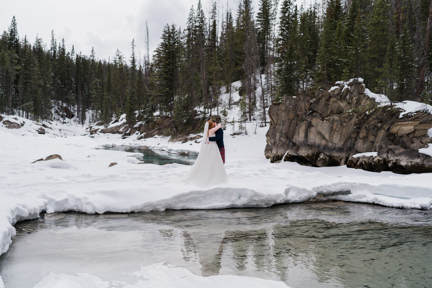 A bride and groom embrace in the snow with their reflection in the river during Emerald Lake elopement