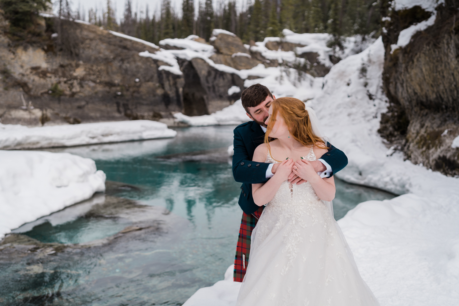 bride and groom embrace with him behind her with bright blue water behind them of the natural bride in yoho national park