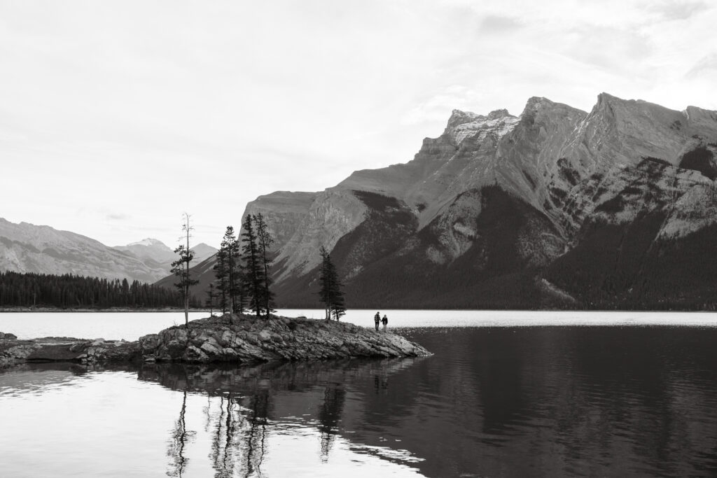 Black and white of a small couple on a rock with trees beside them at Lake Minnewanka after a proposal.