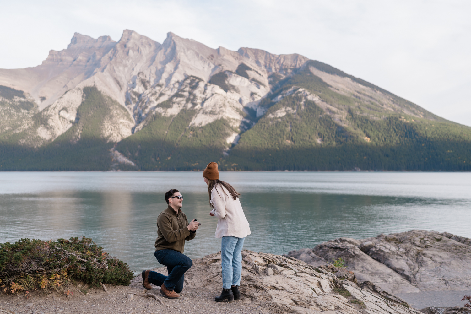 Man kneels infront of surprised girl on the shores of Lake Minnewanka in Banff National Park while proposing. 
