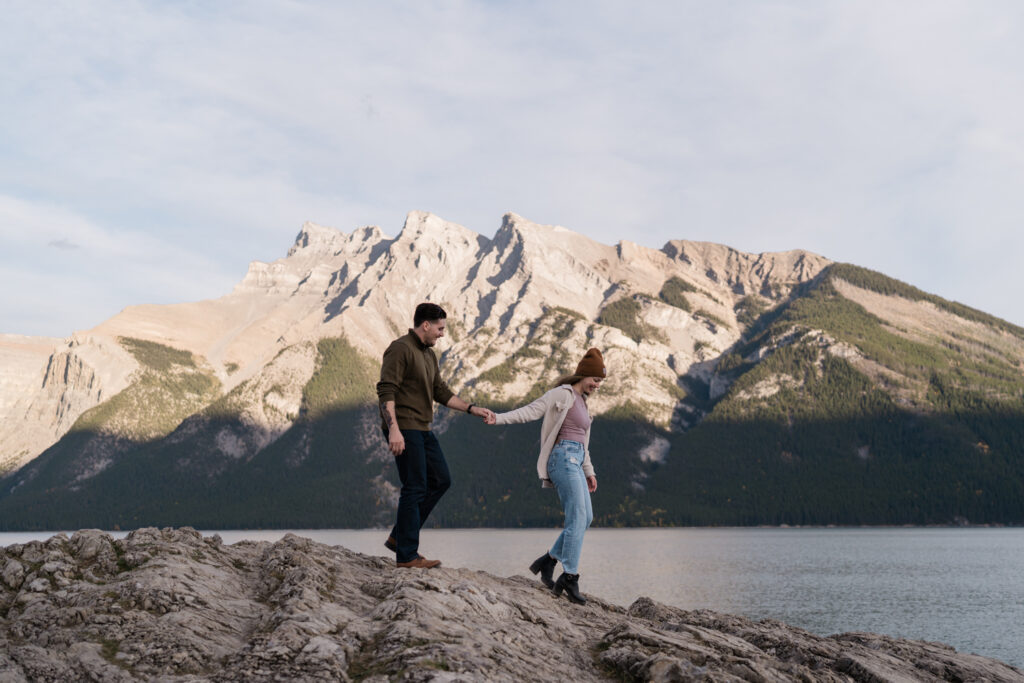 woman leads a man by hand down a rock in front of Lake Minnewanka at sunset