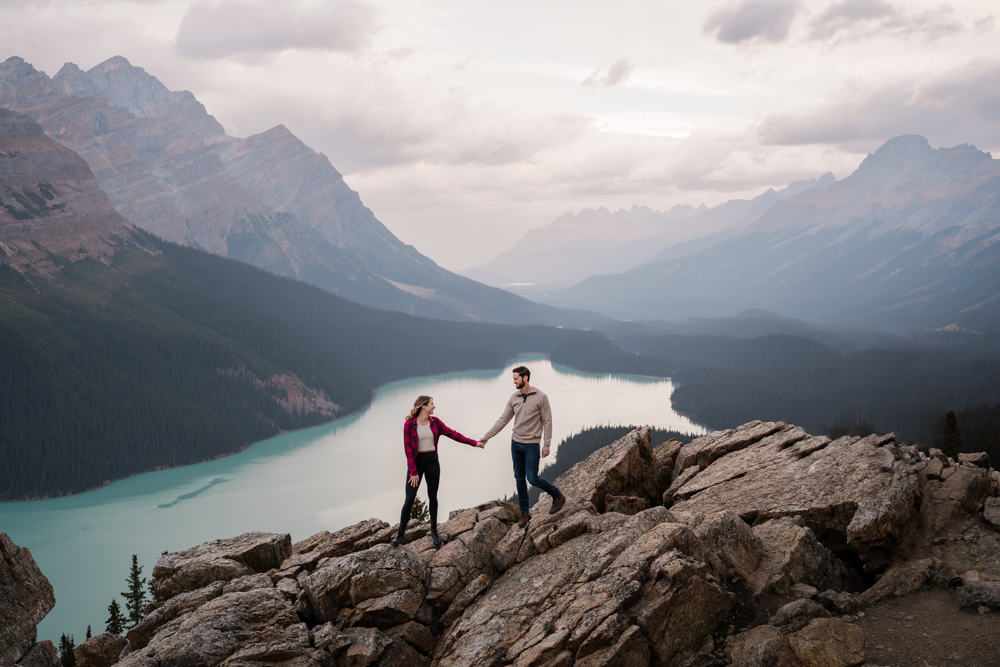 woman in plaid looks back at new fiance as they stand on the rocks above Peyto Lake during sunrise during a Banff National Park engagement session