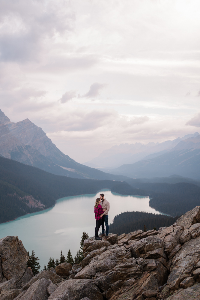 couple stands on rocks above Peyto Lake while they cuddle and he kisses her forehead.