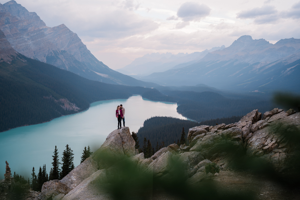 trees in the foreground and a couple stands on a tall rock in front of Peyto Lake at sunrise