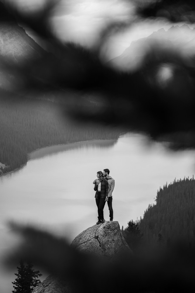 black and white photo of a couple perched on a rock overlooking Peyto Lake with pine tree branches framing them