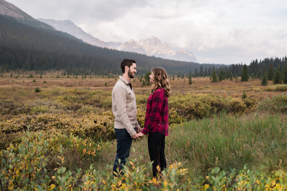 couple holds hands and looks at each other while surrounded by a meadow and mountains during a Banff Engagament session