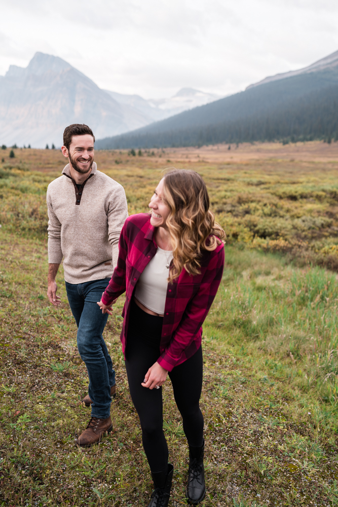 Couple walks in meadow in Banff National Park and she laughs out of focus in front of him