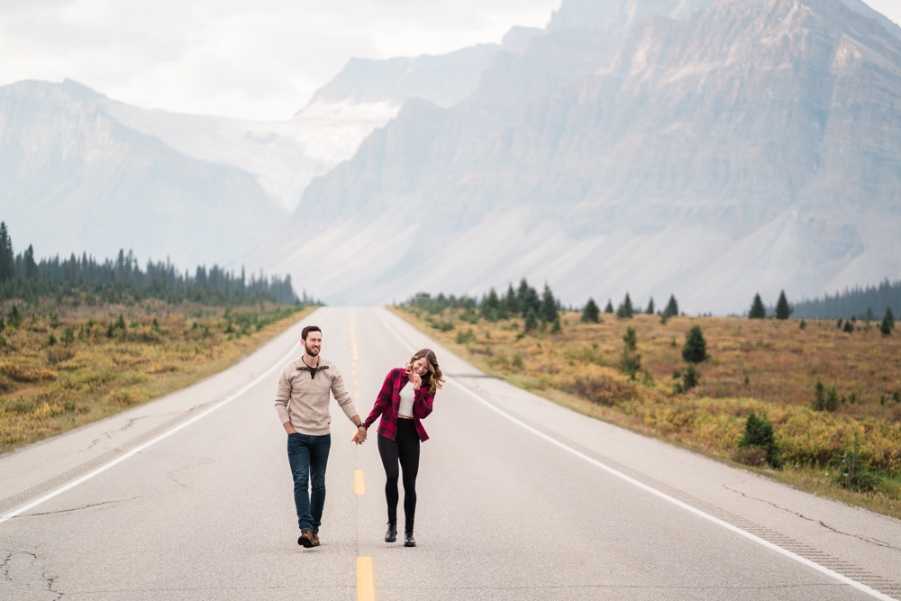 couple walks along the center line with the mountains of Banff National Park behind them while they hold hands and laugh
