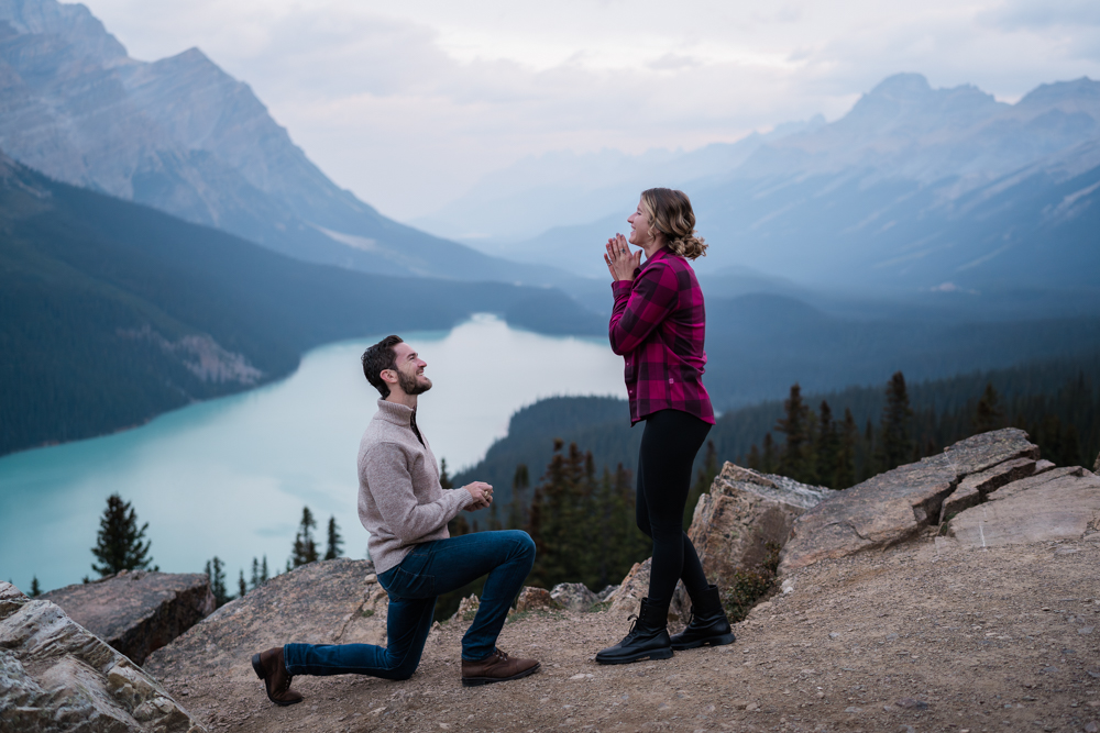 man on one knee smiles up at woman holding her hands in front of her face as he proposes at Peyto Lake