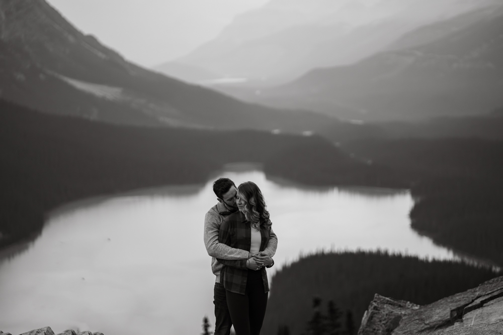 Black and white photo of a man cuddling behind a woman with the wolf shaped Peyto Lake framing them