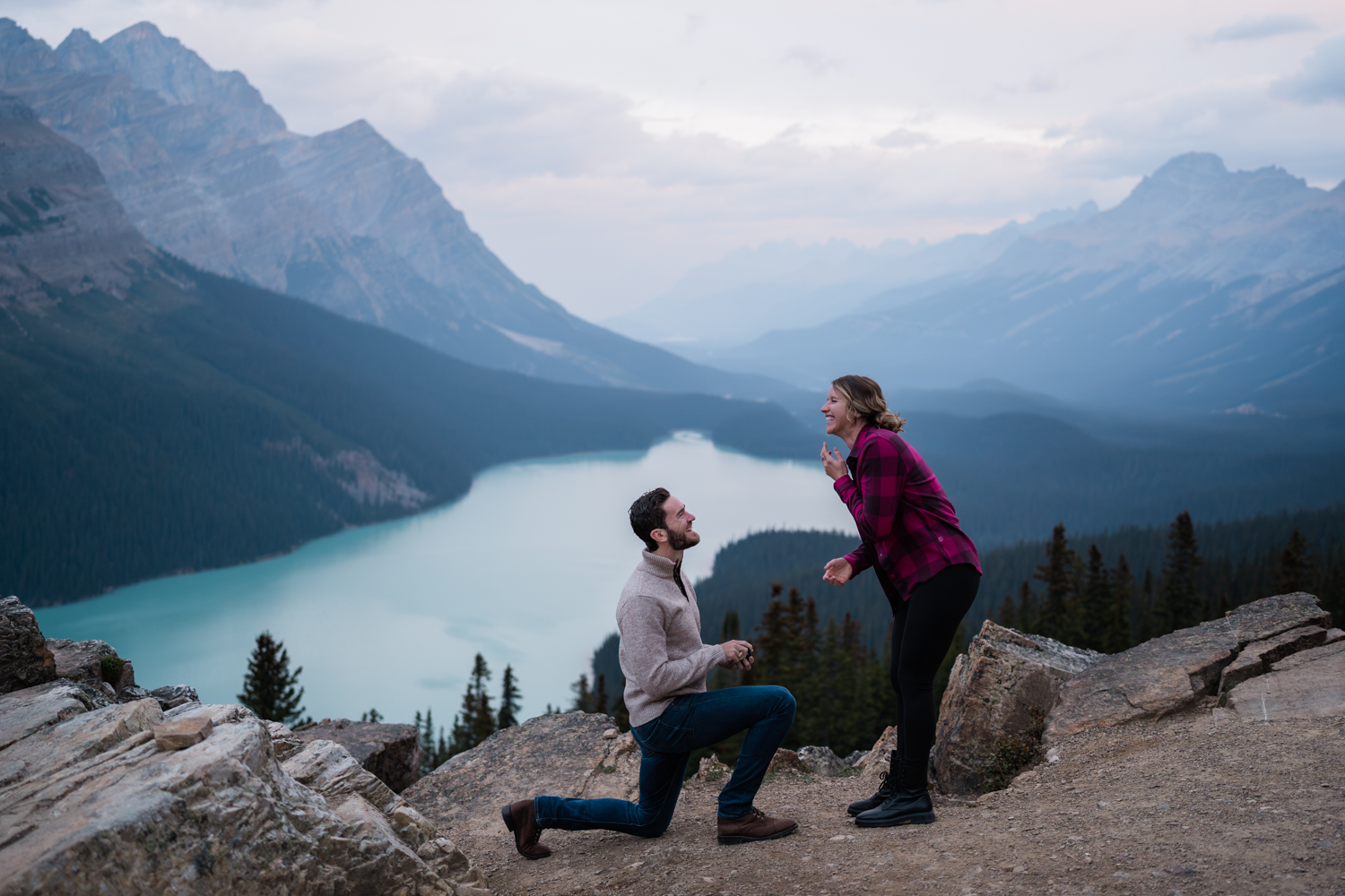 man in grey sweater kneels infront of girl in red plaid shirts during a sunrise proposal at Peyto Lake. 