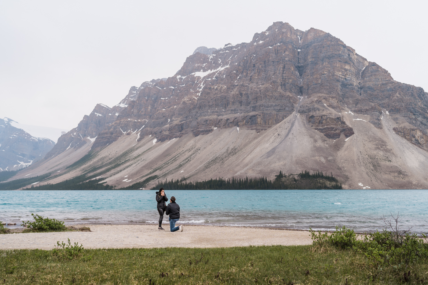 man in black jacket is down on one knee infront of woman in black jacket standing next to Bow Lake in Banff National Park proposing