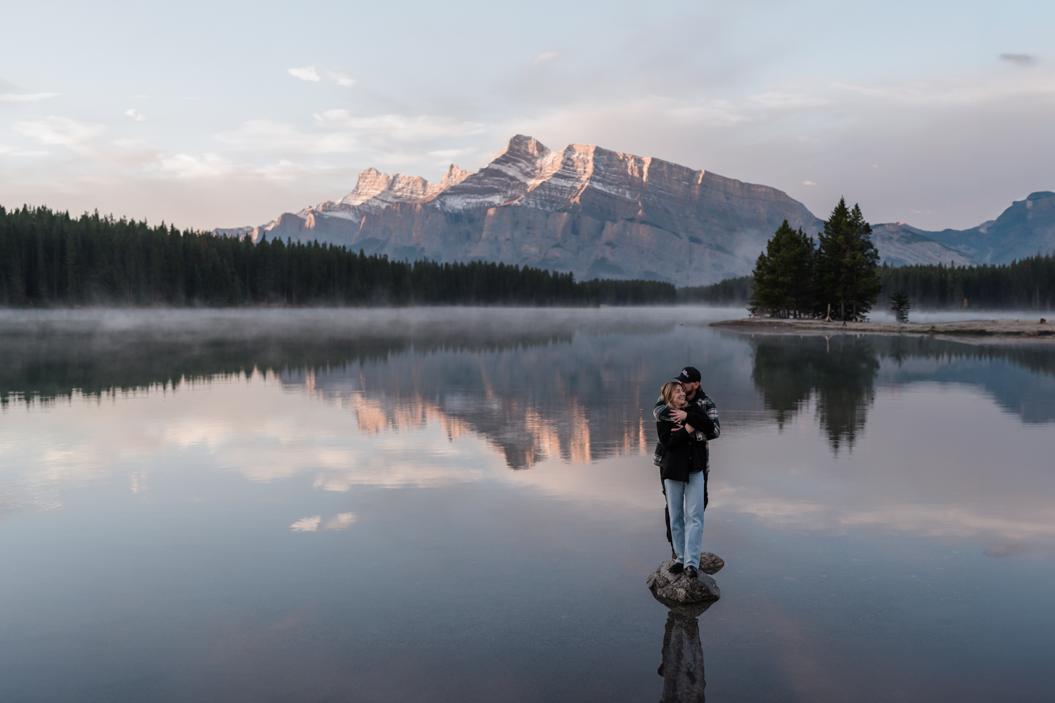 Couple stands cuddling on a rock after a proposal at Two Jack Lake with the lake reflection all around them. 