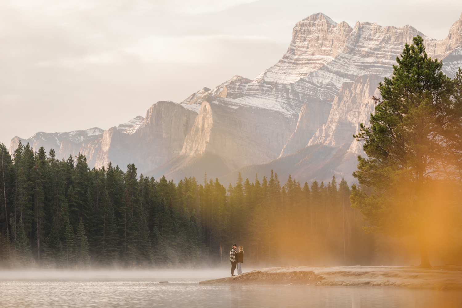Small couple stands on side of Two Jack Lake during sunrise with yellow leaves in the foreground and the mountain lit up behind them. 