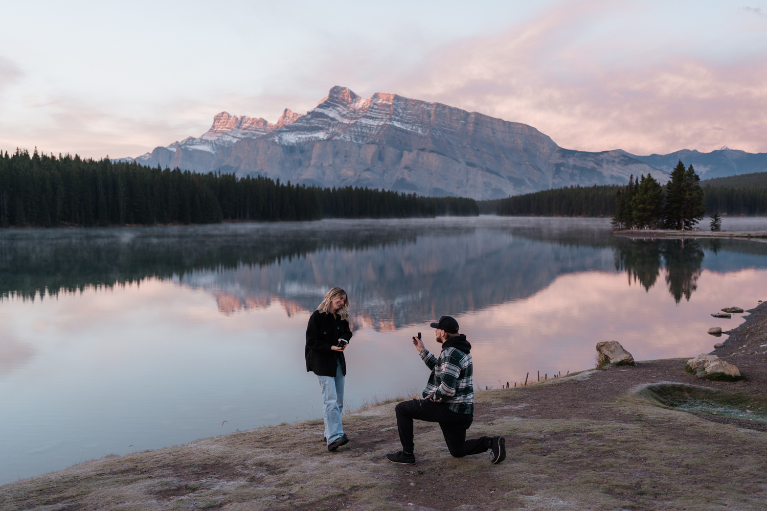 Man in plaid is on one knee infront of woman in black during sunrise proposal at Two Jack Lake in Banff National Park