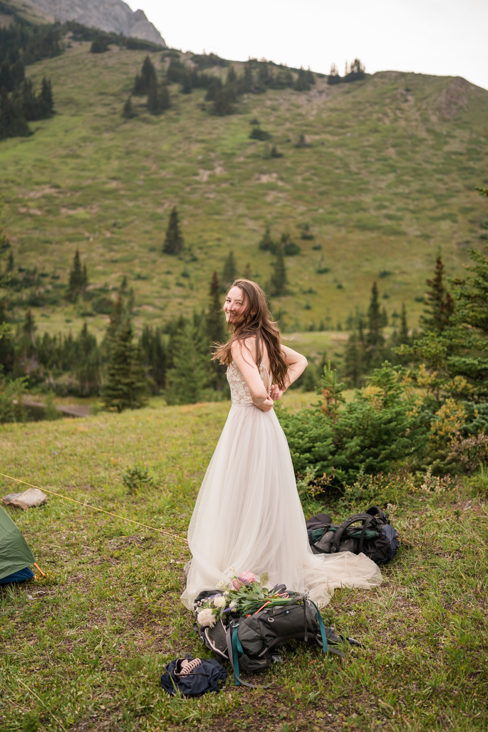 Bride does up her dress in the meadow during camping elopement.