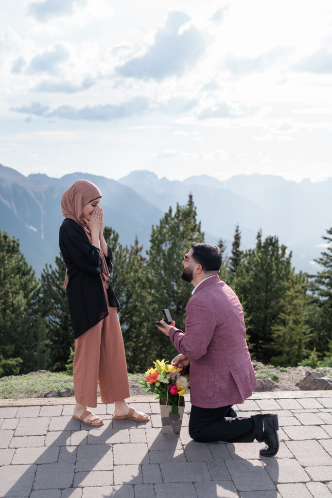 man kneels infront of a girl with her hands over her face proposing in the summer on top of the Banff Gondola. 