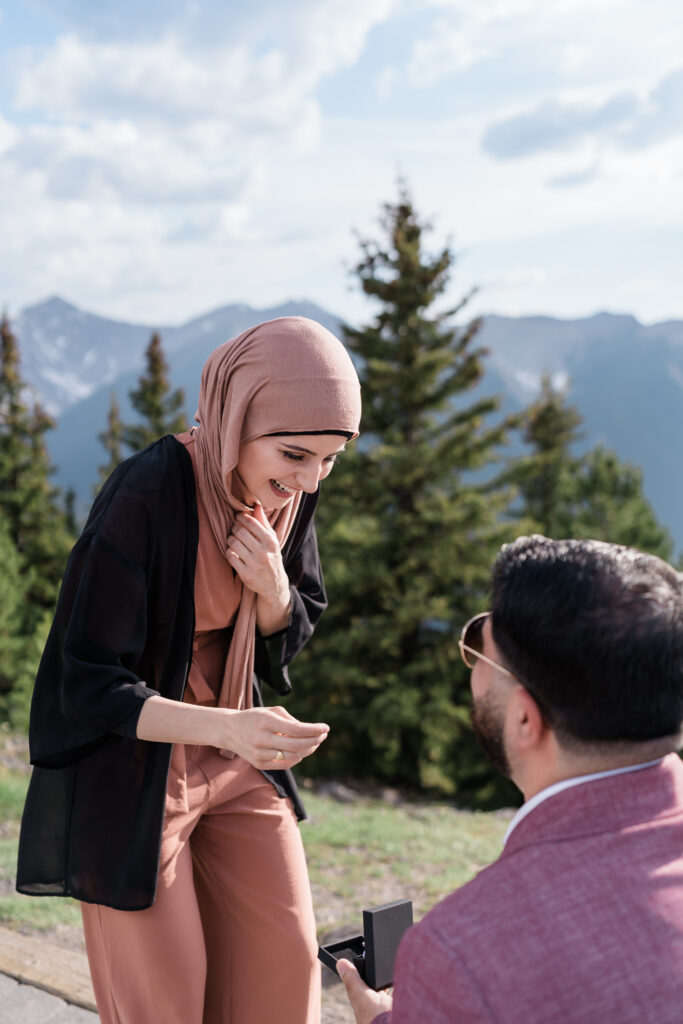 girl in pink head scarf looks down at man kneeling and holding a ring box and is surprised and happy after proposal on the Banff Gondola