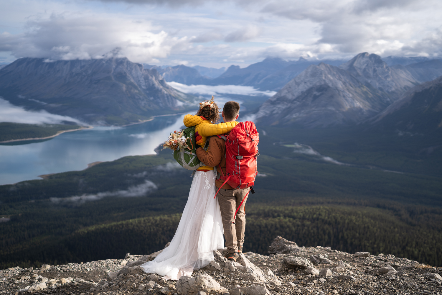 bride in yellow jacket with flowers in her backpack cuddles with groom with red backpack on tent ridge during hiking elopement in kananaskis