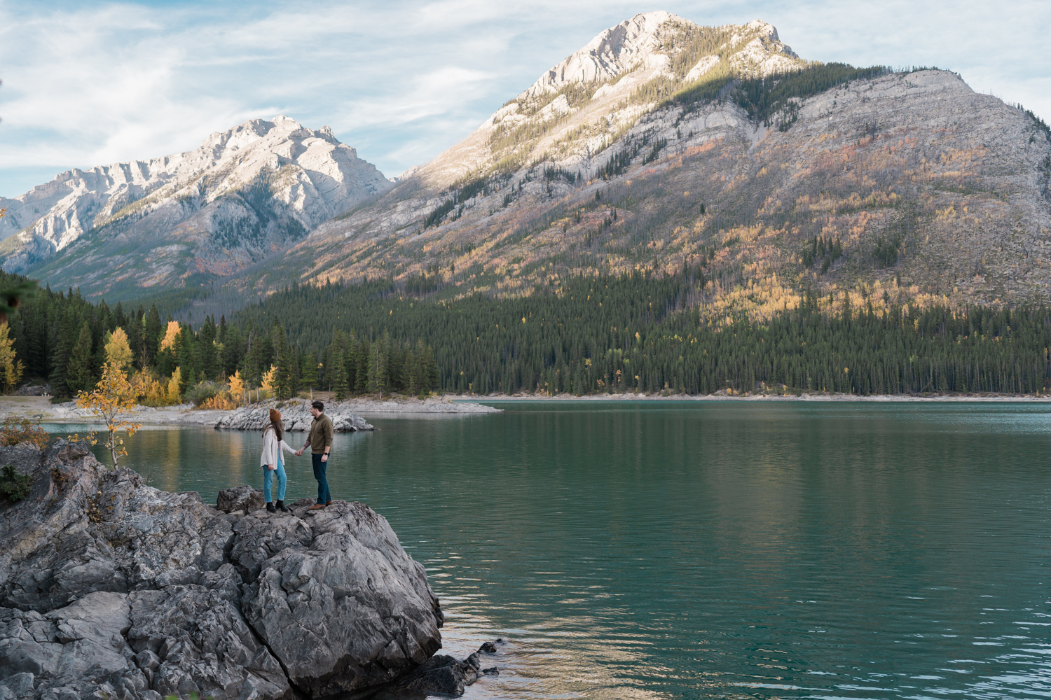 Couple holds hands and looks over at the fall colours in Banff at Lake Minnewanka. 