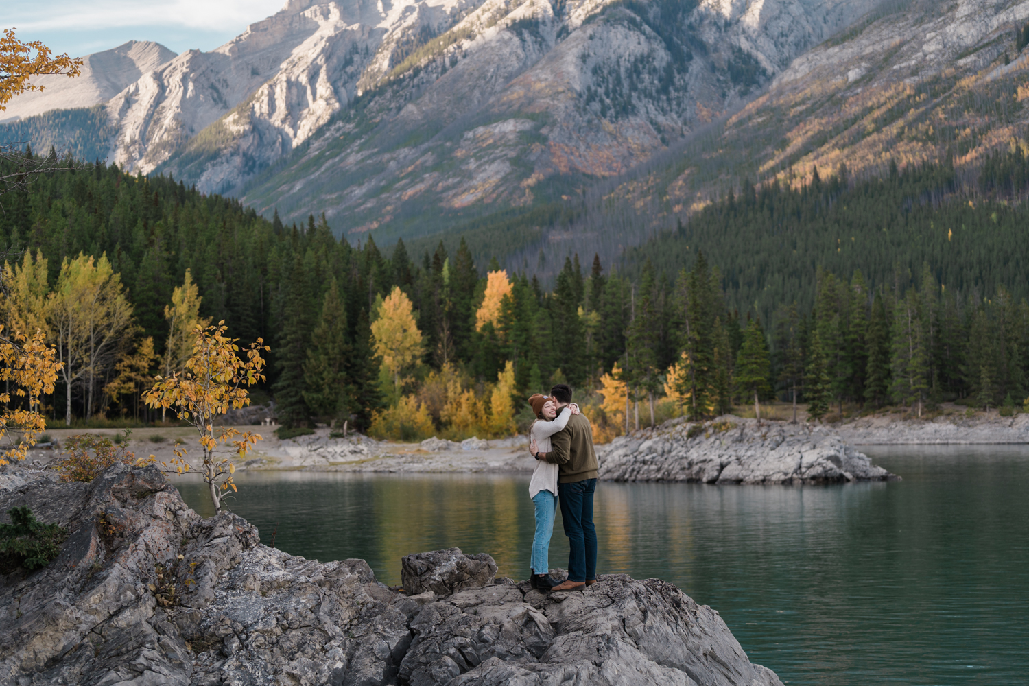 Couple holds each other in front of the yellow leaves and lake. 