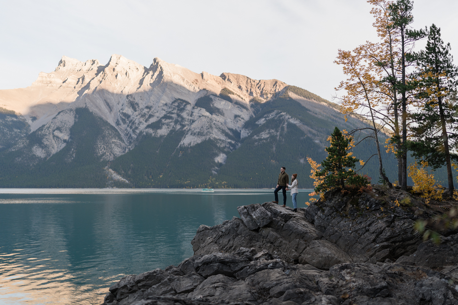 Couple stands holding hands overlooking Lake Minnewanka after Banff proposal. 