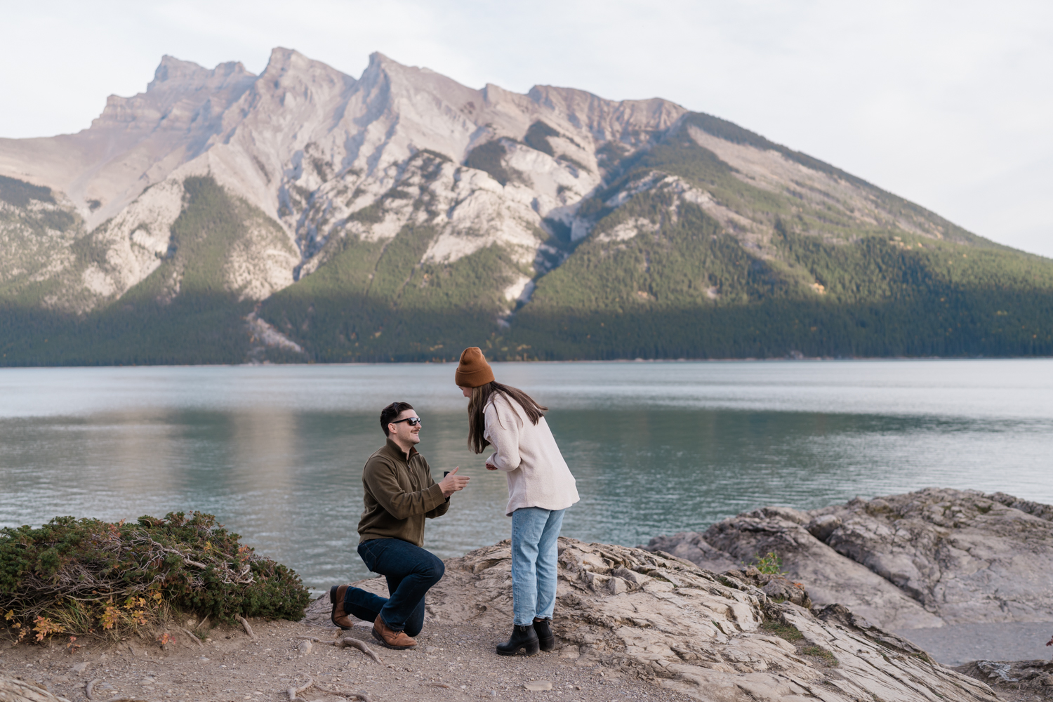 Man kneels in front of his girlfriend and holds a ring box up to her by Lake Minnewanka in Banff. 