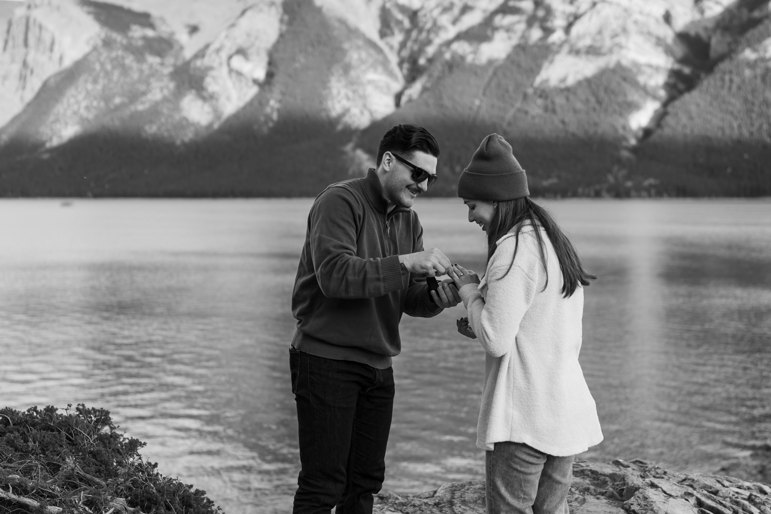 Man slides ring on woman's finger during Lake Minnewanka proposal.