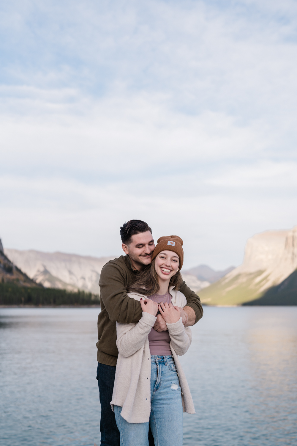 Woman smiles while fiance cuddles her from behind in front of Lake Minnewanka. 