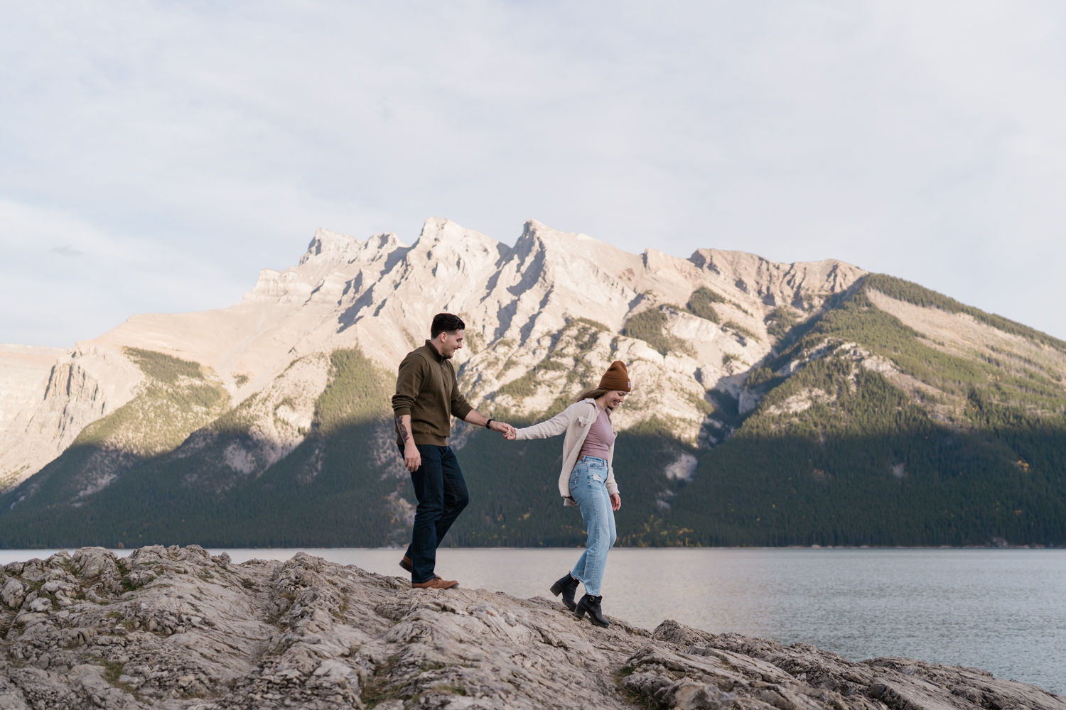 Couple holds hands while walking by Lake Minnewanka for Banff engagement photos. 